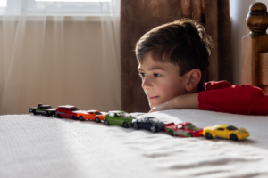 A boy in a red shirt lines up toy cars on his bed